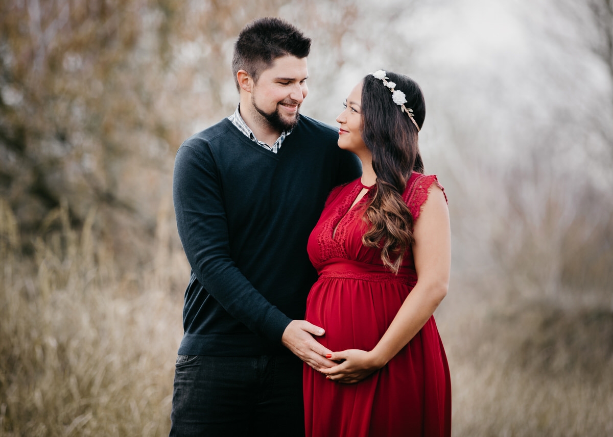 Mann steht mit seiner schwangeren Frau, deren Haar von einem Blumenkranz gekrönt werden, im Arm vor einem lichten Wäldchen. Beide haben eine Hand auf ihrem Bauch liegen sie blicken sich an und lächeln verliebt.
 Fotografin: Claudia Nürnberger