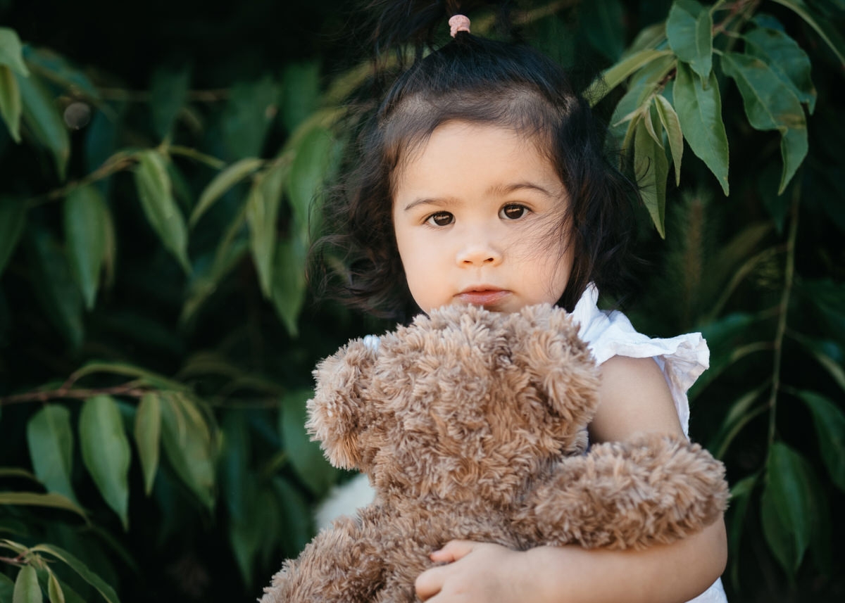 Kleines Mädchen hält einen Teddy in ihrem Arm und schaut etwas schüchtern mit großen Augen direkt in die Kamera. Fotografin: Claudia Nürnberger