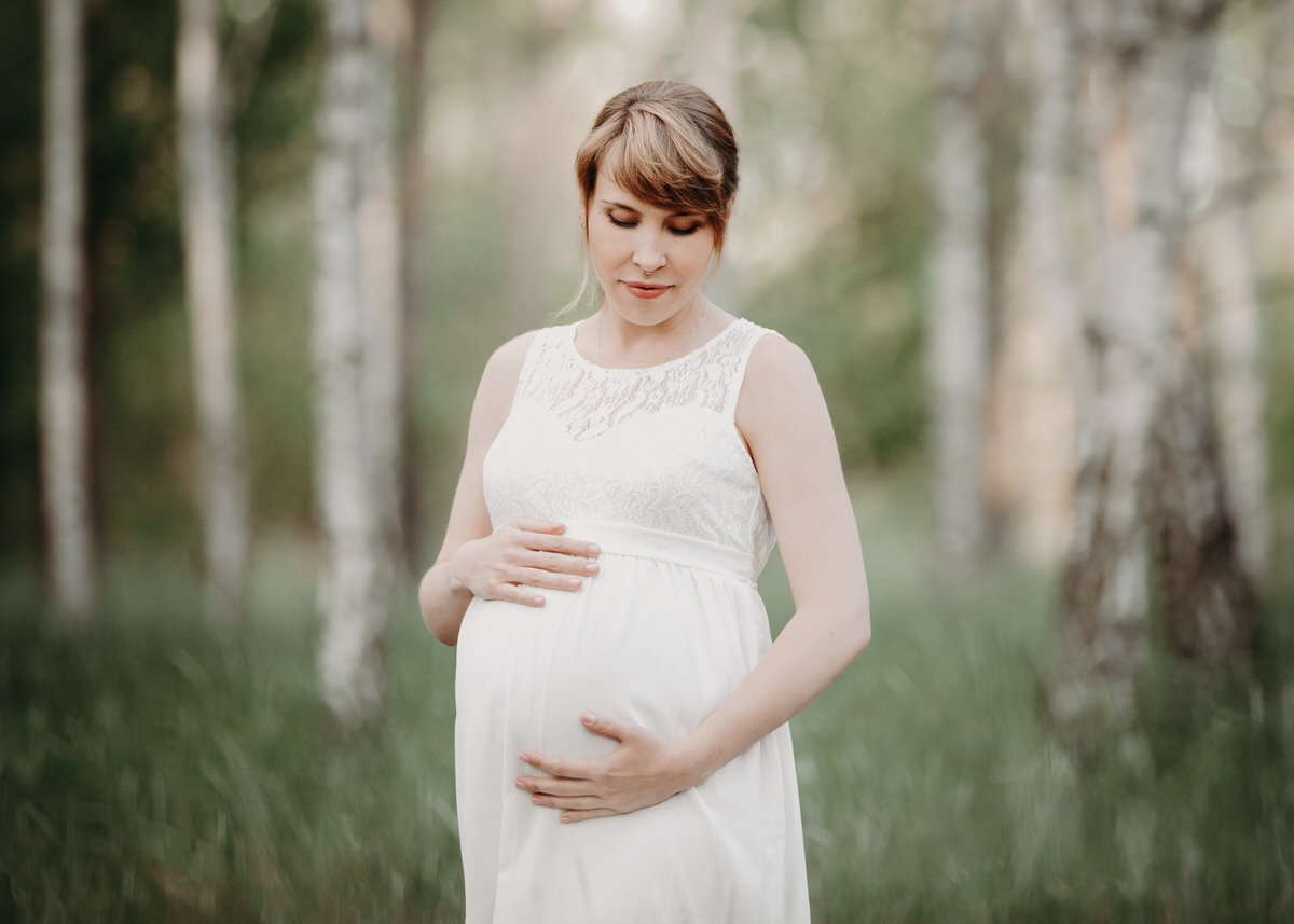 Schwangere Frau steht vor einem Hintergrund aus Birken in der Natur und schaut liebevoll auf ihren Babybauch
 Fotografin: Claudia Nürnberger