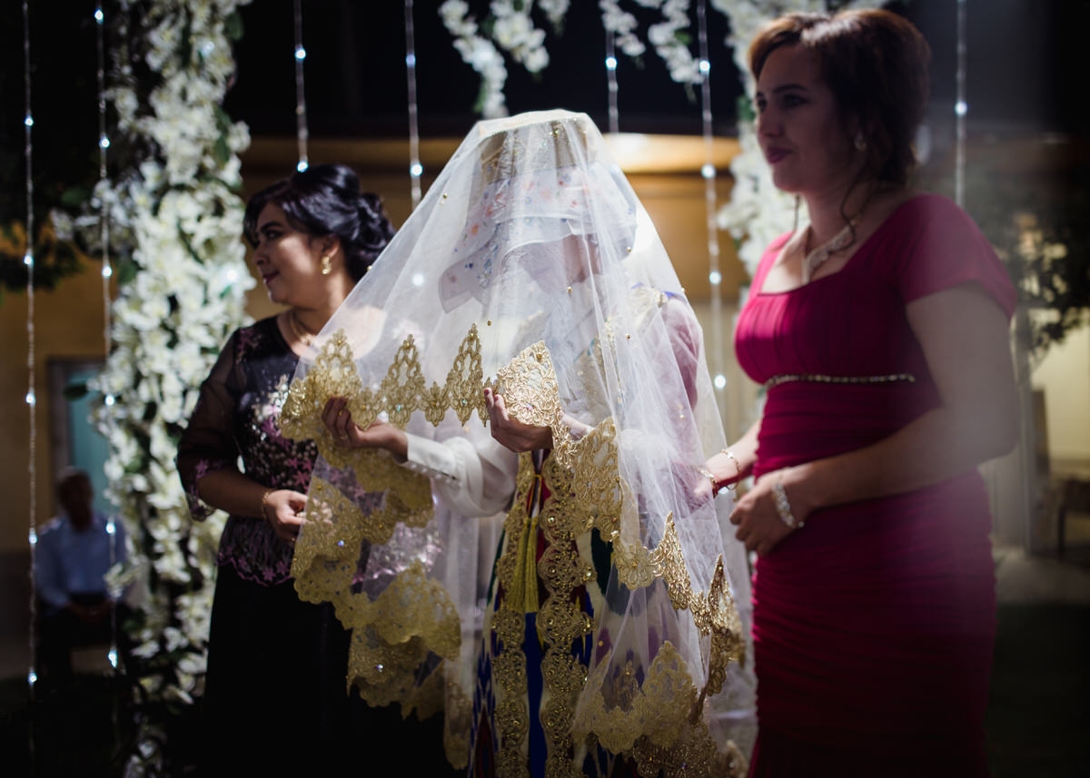Braut steht unter einem geschmückten Pavillon in einer aufwendigen Traditionellen Hochzeitstracht aus Usbekistan. Ihr Kopf ist gesenkt und sie hält ihren Schleier mit beiden Händen fest. Fotografin: Claudia Nürnberger