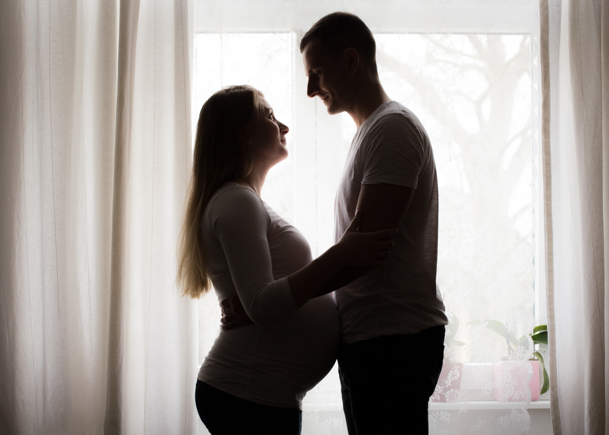 Ein schwangeres Pärchen steht vor einem Fenster im Profil und hat die Gesichter einander zugewandt. Sie umarmen sich zärtlich und ihre Bäuche berührt sich, während sie sich anlächeln.
 Fotografin: Claudia Nürnberger