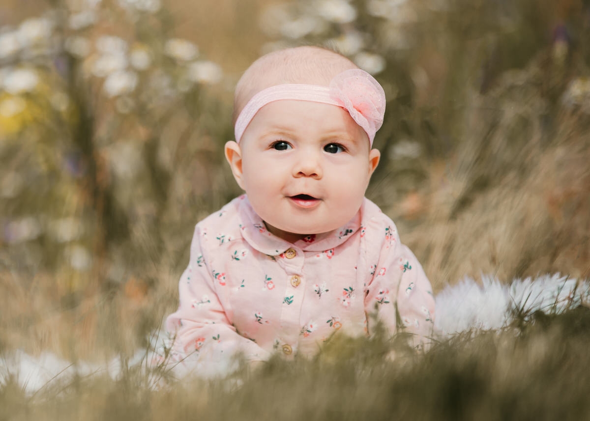 Ein Kleinkind liegt auf einem weißen Fell vor einer Blumenwiese. Sie trägt ein Haarband und schaut lächelnd nach links. Fotografin: Claudia Nürnberger