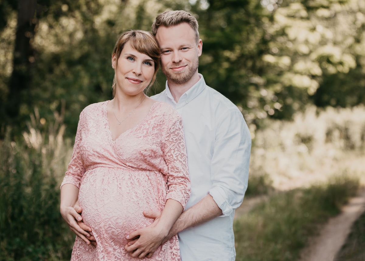 Mann steht hinter seiner schwangeren Frau auf einem Feldweg. Ihre Hände liegen aufeinander auf ihrem Babybauch während beide glücklich dem Betrachter zugewendet lächeln.
 Fotografin: Claudia Nürnberger