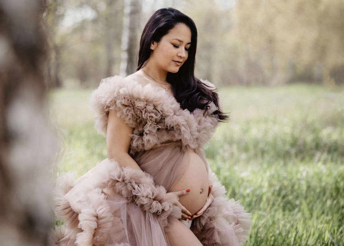 Schwangere Frau, steht in der Natur und schaut glücklich auf ihren Babybauch. Ihr ausladendes mit Rüschen bedecktes Kleid umschließt ihre Schultern und wallt über ihre Seiten bis zum knöchelhohen Gras.
 Fotografin: Claudia Nürnberger
