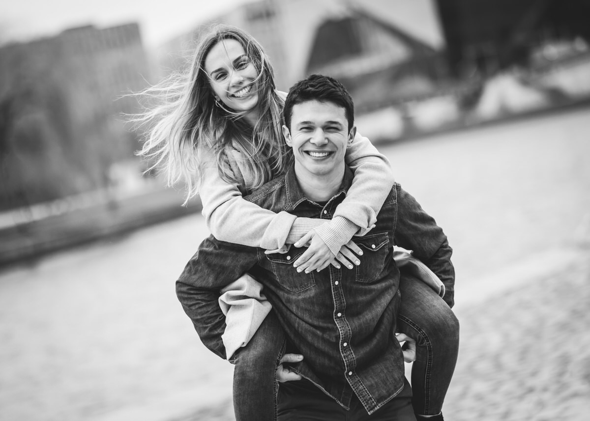Junger Mann trägt seine Freundin Huckepack. Beide lachen und ihre Haare fliegen im Wind. Fotografin: Claudia Nürnberger