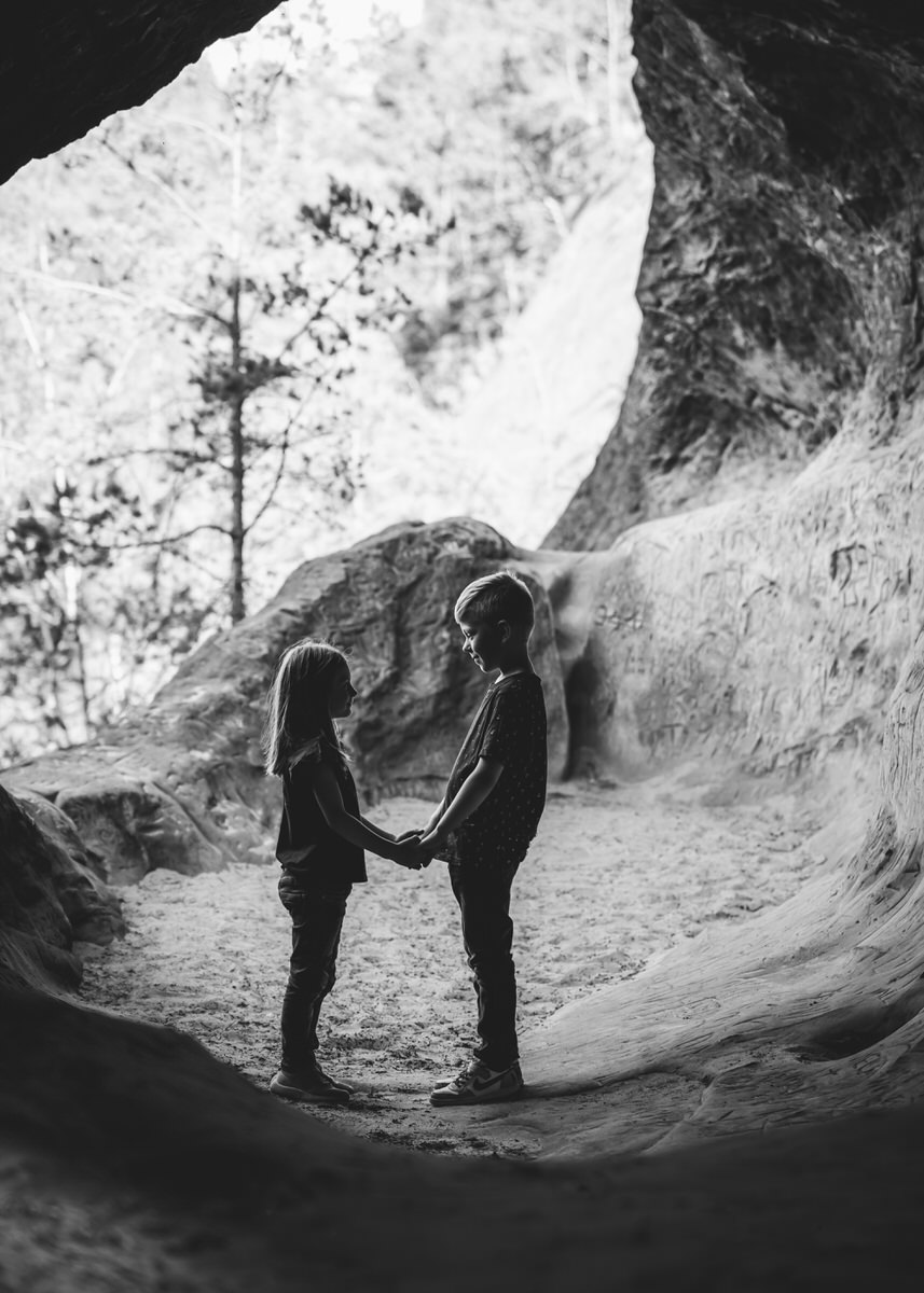 Ein Geschwisterpaar steht in einer Höhle. Im Hintergrund sieht man den Ausgang der Höhle. Beide stehen sich gegenüber und halten sich gegenseitig ihre Hände. Sie lächeln und blicken sich direkt an. Fotografin: Claudia Nürnberger