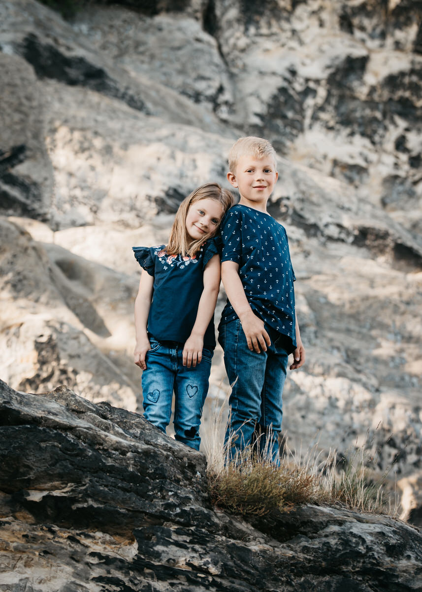 Ein Junge und ein kleineres Mädchen stehen auf einem Felsen Rücken an Rücken und schauen runter auf den Betrachter und lächeln. Fotografin: Claudia Nürnberger