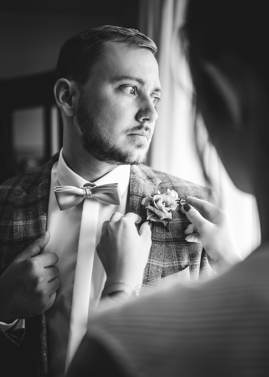 Braut richtet die Blume am Reviere des Bräutigams. Er blickt hinaus auf dem Fenster vor den die beiden stehen.Fotografin: Claudia Nürnberger / Berlin