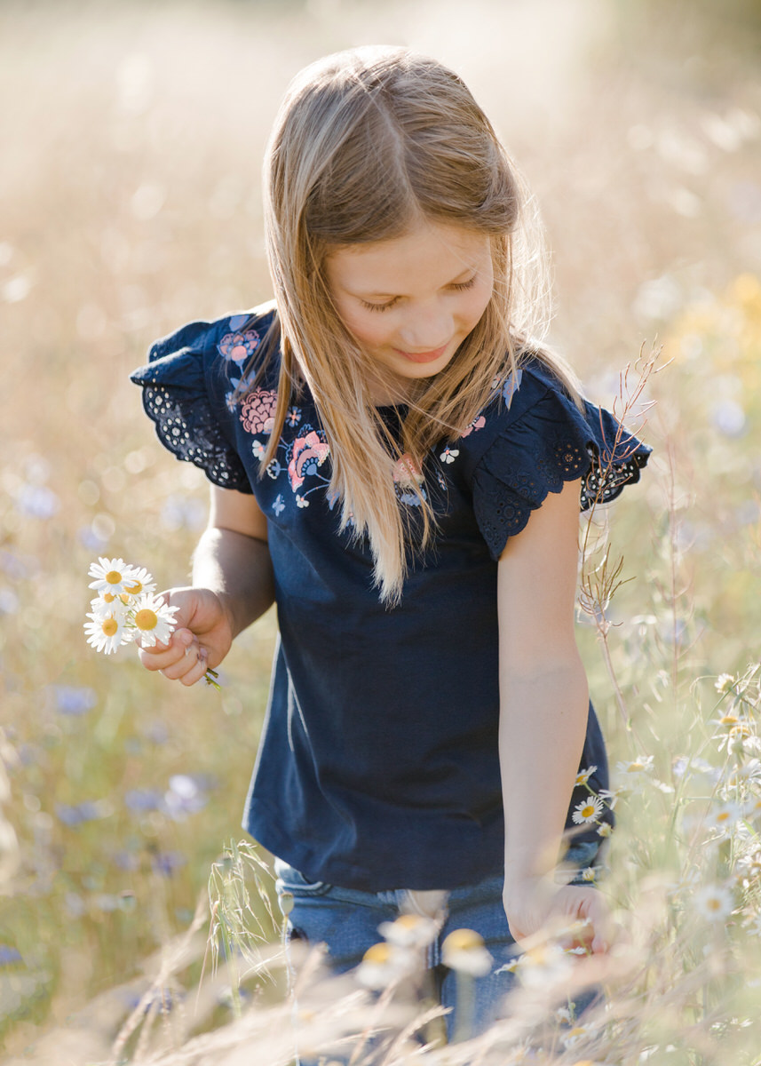 Ein Mädchen im Grundschulalter steht in einer Blumenwiese und pflückt grade einen kleinen Blumenstrauß. Sie schaut zu den Blumen herunter und lächelt. Fotografin: Claudia Nürnberger