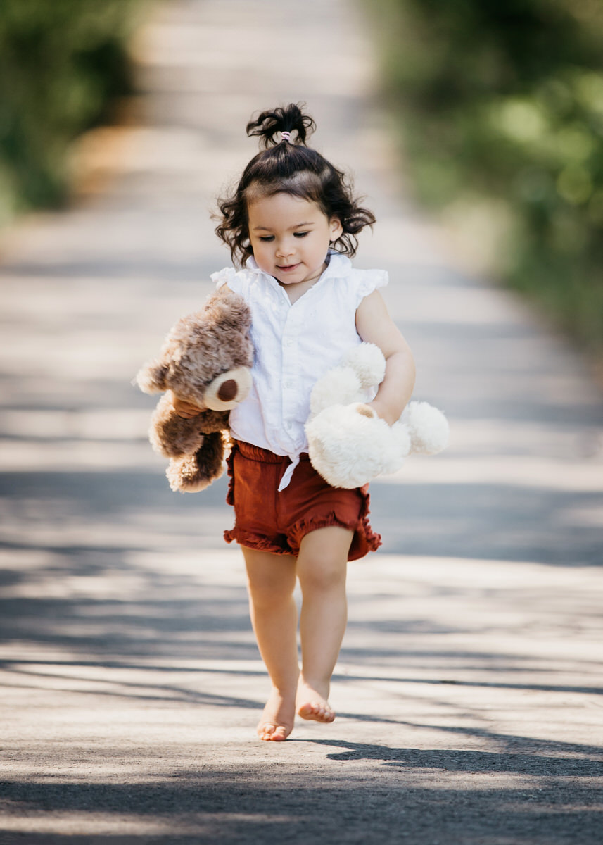 Kleines Mädchen rennt einen Weg entlang direkt zum Betrachter hin. Unter jedem Arm hat sie einen Teddybären. Ihr Blick geht lächelnd gen Boden. Fotografin: Claudia Nürnberger