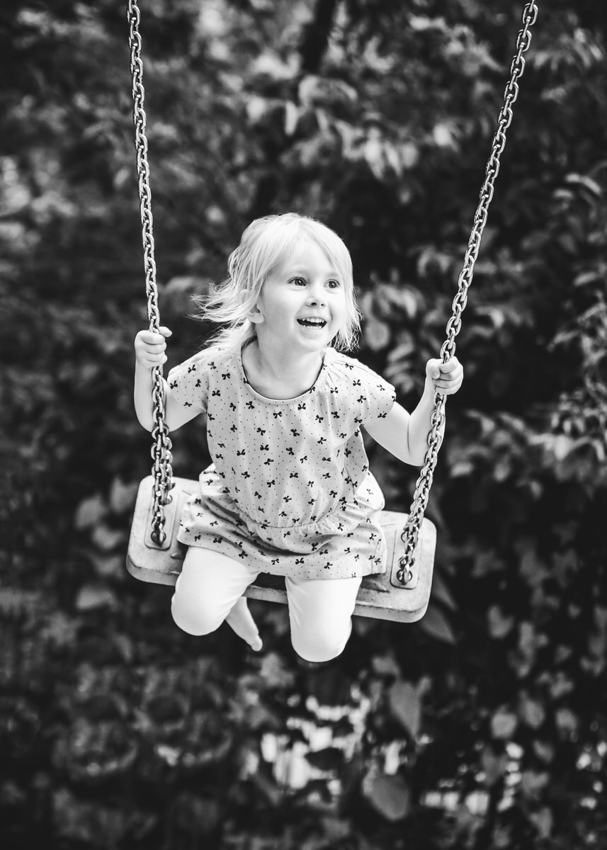 Kleines Mädchen schaukelt wild auf einer Schaukel. Sie lacht herzlich und guckt rechts am Betrachter vorbei. Ihre Haare fliege im Wind. Fotografin: Claudia Nürnberger
