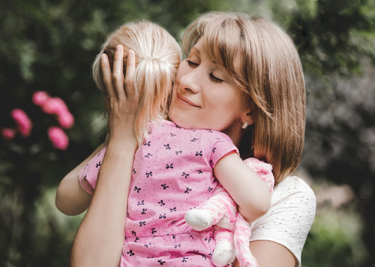 Mutter umarmt ihre Tochter. Ihre Hand hält den Kopf des kleinen Mädchens. Die Augen der Frau sind geschlossen und sie lächelt glücklich. Fotografin: Claudia Nürnberger