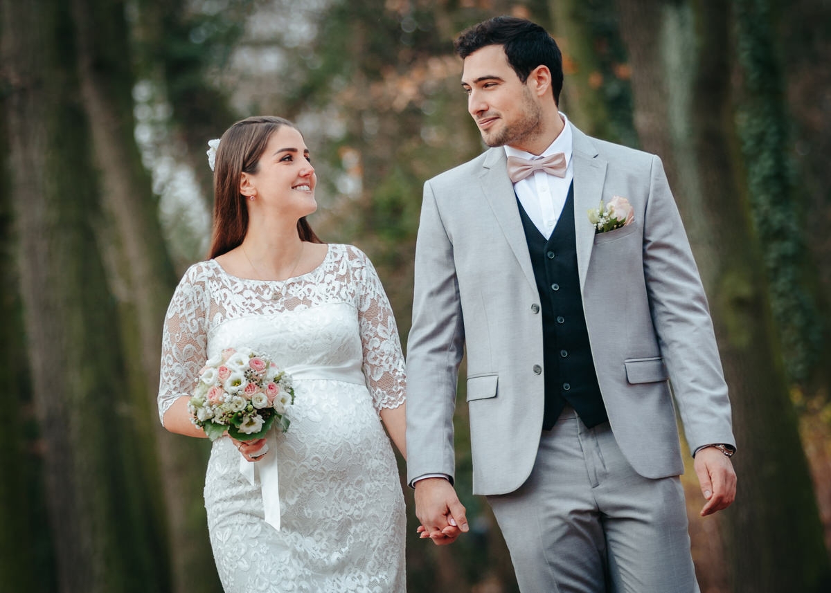 Schwangere Braut und ihr Mann laufen Hand in Hand auf den Betrachter zu. Beide schauen sich an und lächeln verliebt. Fotografin: Claudia Nürnberger